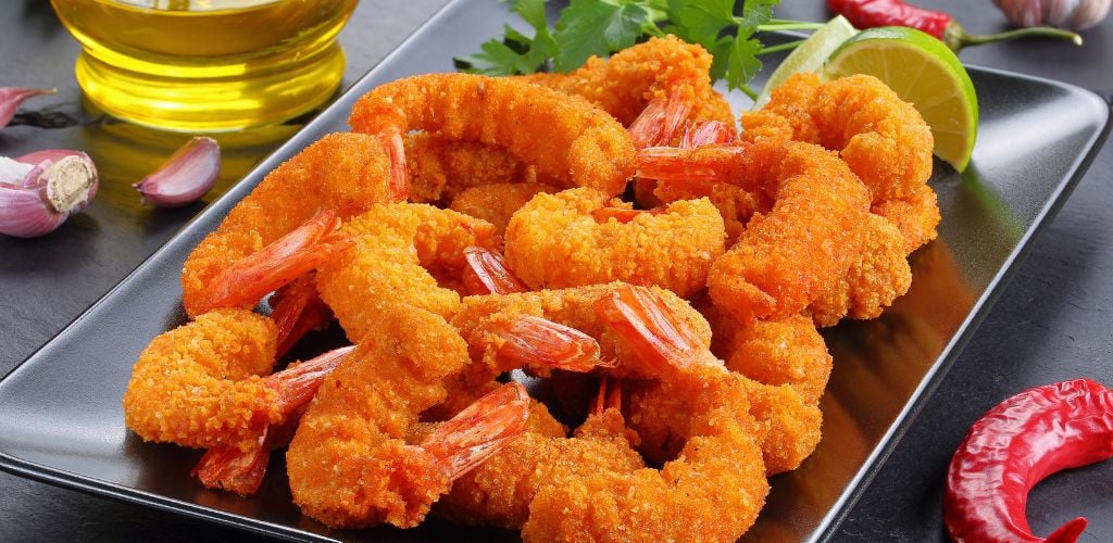 deep-fried breaded shrimp on a plate with lime, red chili, and green leaf on the side.