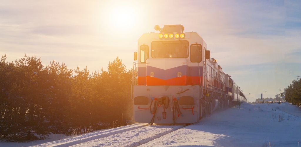 Is a train with snow on the tracks and trees on the side against the backdrop of a sunset. 