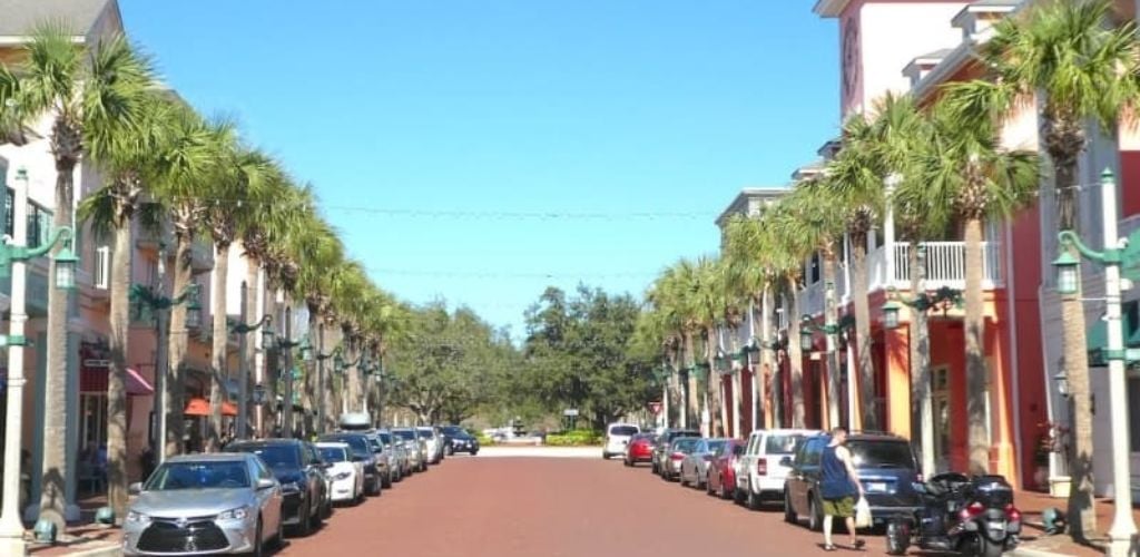 A street with parked cars on the sidewalk, trees, and buildings, and a man walking with a white plastic bag. 