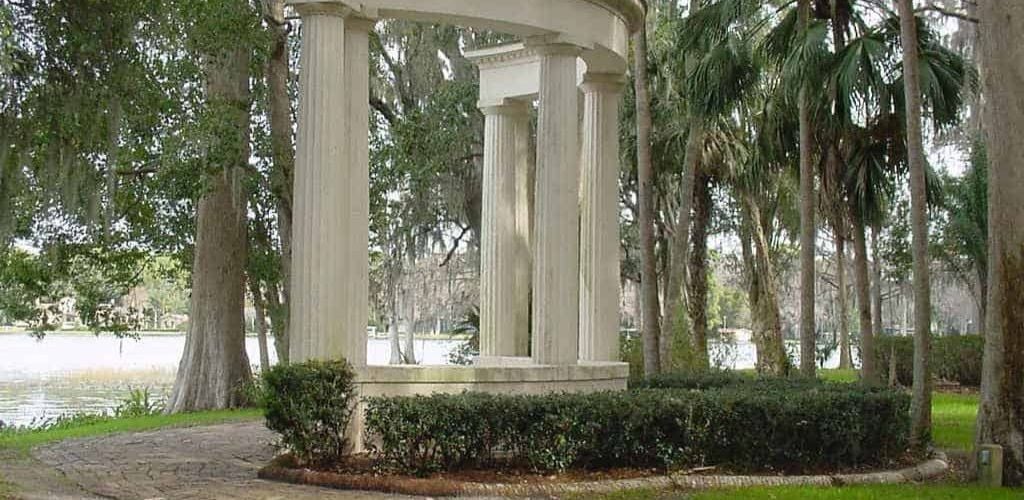 A Greek Architecture style in the front of a lake, surrounded by green plants and trees. 