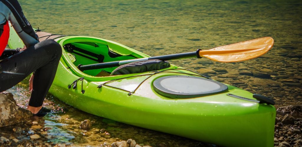 At the bank lake, there is a green kayak park and a yellow paddle with a man sitting on the side. 