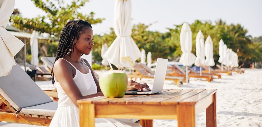 Pretty young woman sitting at a table on a sandy beach, enjoying a fresh coconut cocktail and working on a laptop