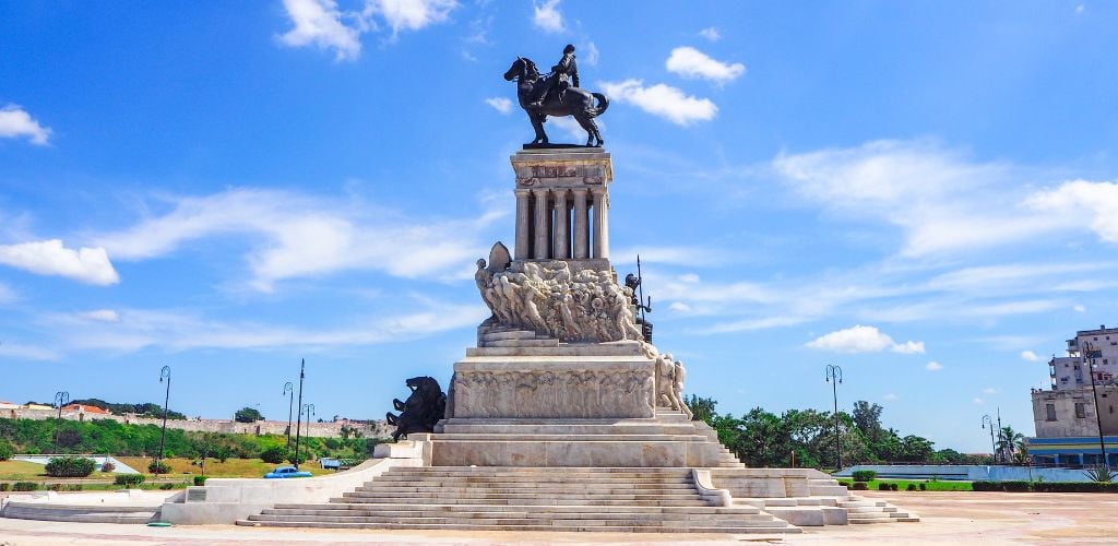 On a sunny day, a statue in the middle of the park with street lights, some building structure, and green trees. 