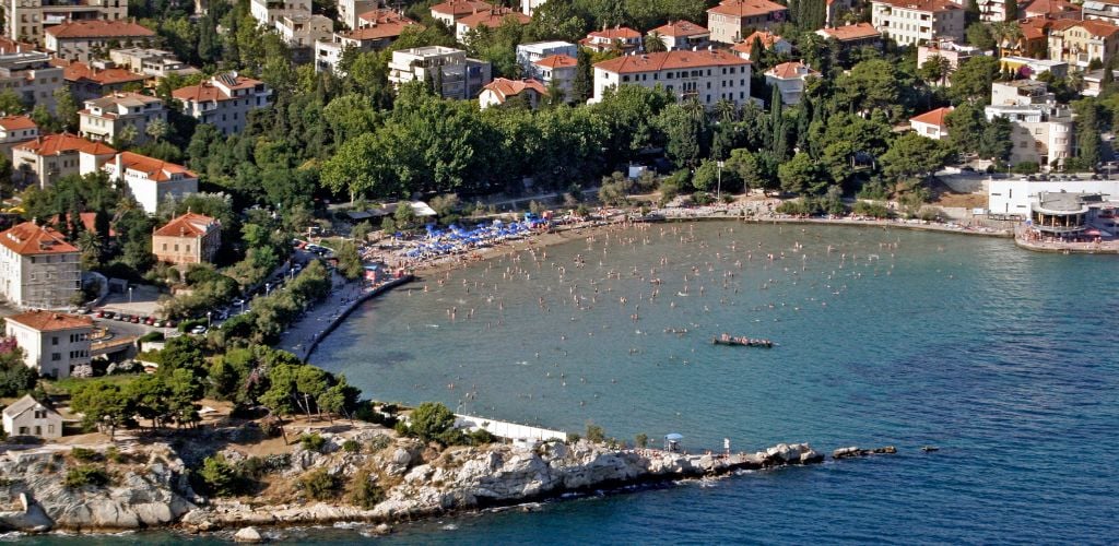 An aerial view of a beach with a large building close to the shore and a large number of tourists on the shoreline, all surrounded by green trees. 