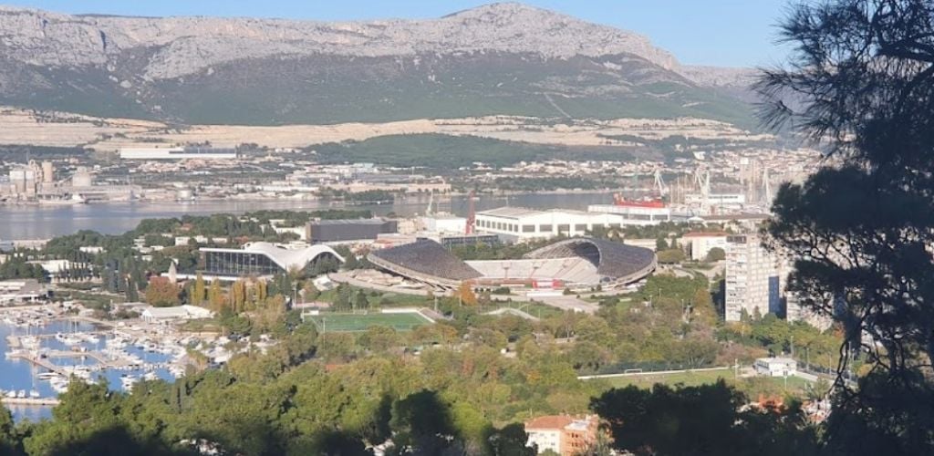 An uphill overlooking a soccer filed, a large pier, and a large mountain in the distance. 