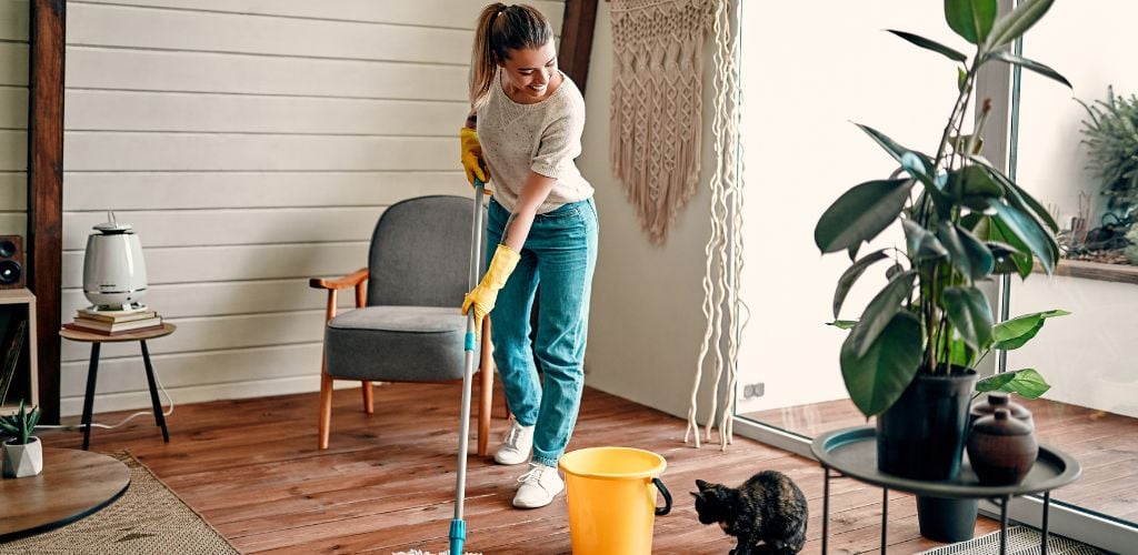 A young attractive girl in rubber gloves and headphones listens to music and mops the wooden floor in the room.