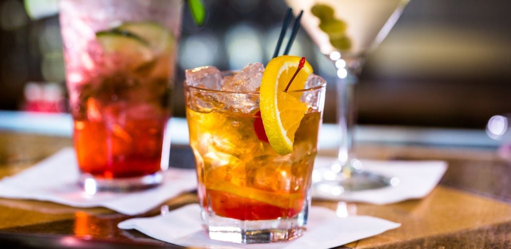 fruity cocktail with ice in a small glass tumbler with a slice of lemon and a cherry, colorful cocktails in background of image