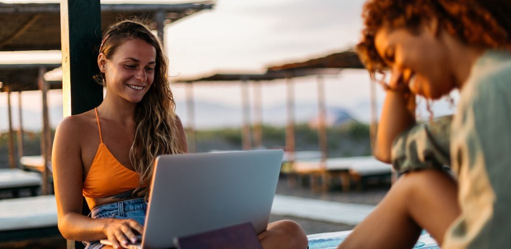 digital nomad friends with laptop working from a beach location laughing and smiling