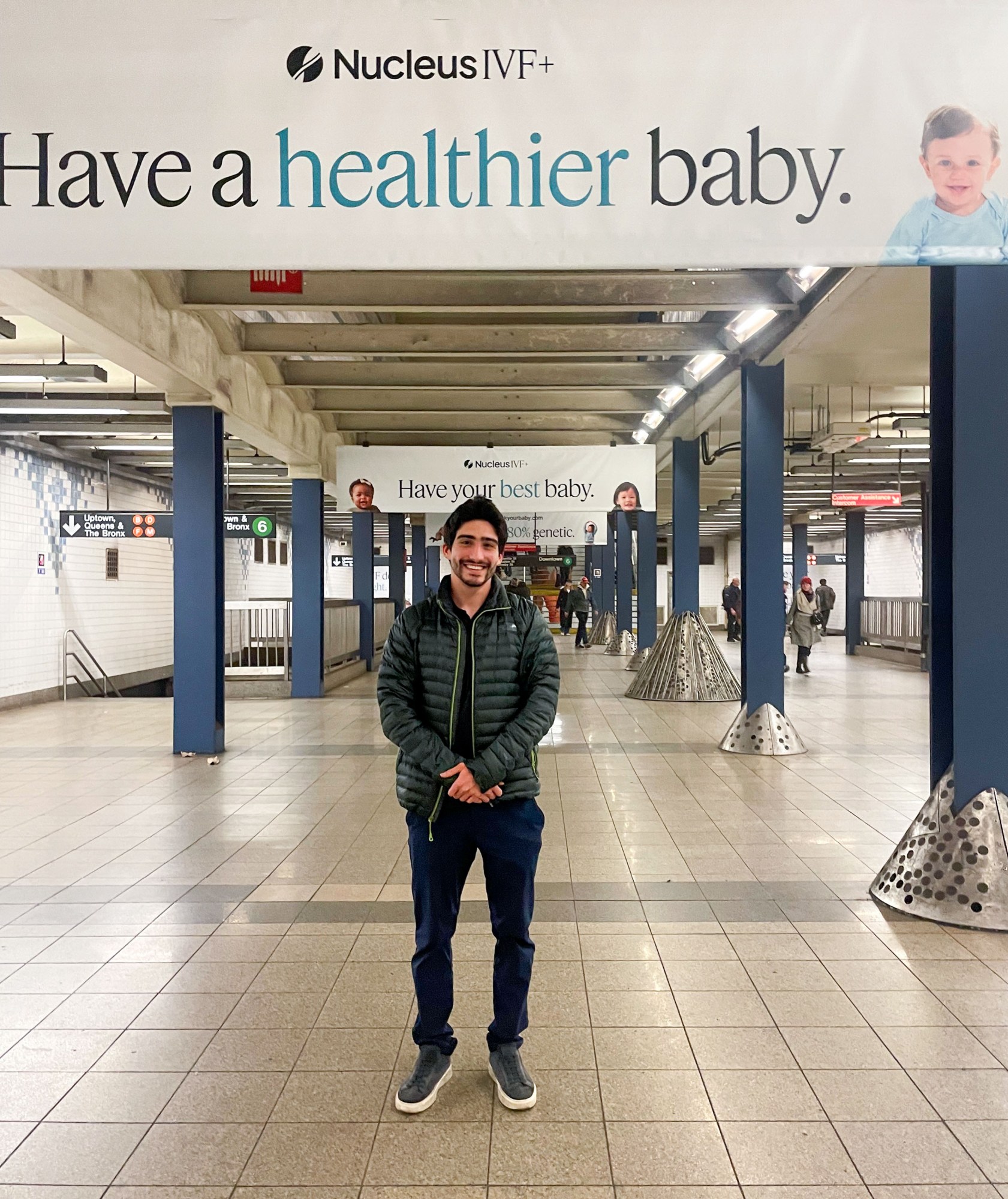 Sadeghi poses under the first in a row of advertisements. The one above him reads, "Nucleus IVF+ Have a healthier baby." with the word "healthier" emphasized.