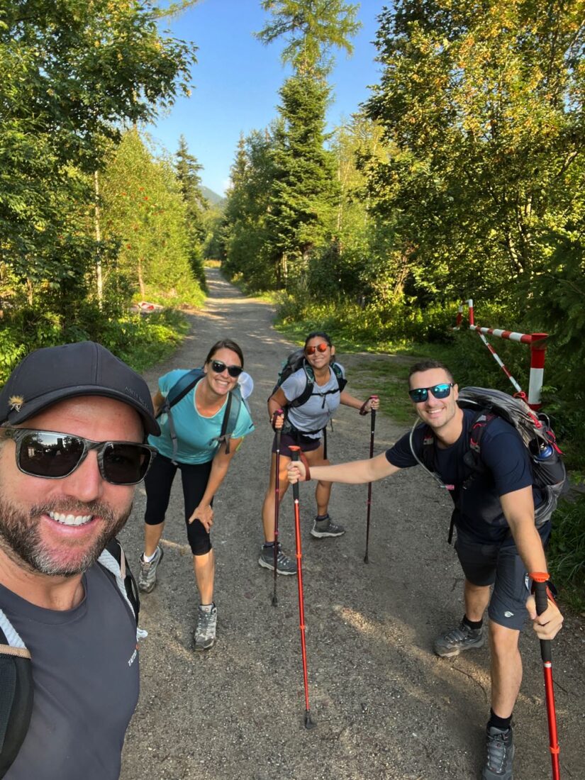 Goats On The Road Nick and Dariece hiking with friends in outdoor setting, green trees line path and bright blue skies