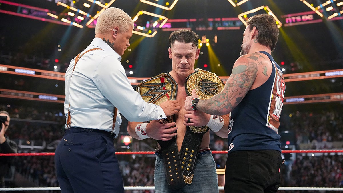 Cody Rhodes and CM Punk handing John Cena two WWE championship belts in an arena